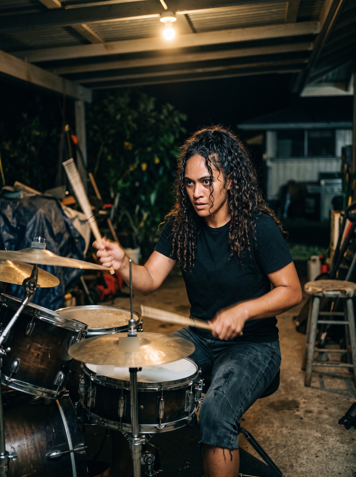 A drummer with long dark curly hair plays hard on a worn kit in a cluttered carport at night under a bare bulb